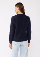 [Color: Navy] A brunette model wearing a classic crew neck sweater in navy blue. With ribbed trims, long sleeves, and a cable knit stitch detail. A versatile sweater perfect for layering during colder temperatures or wearing on its own. 