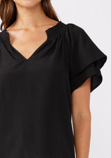 [Color: Black] A brunette model wearing a sophisticated black top. With double short flutter sleeves, smocked neck detail, split v neck, and a relaxed fit. An everyday top perfect for the office or a casual outing.