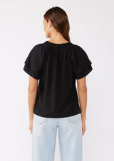 [Color: Black] A brunette model wearing a sophisticated black top. With double short flutter sleeves, smocked neck detail, split v neck, and a relaxed fit. An everyday top perfect for the office or a casual outing.