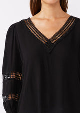 [Color: Black] A brunette model wearing a black casual top with lace trim or crochet inserts. With a v neckline, long sleeve with elastic cuff, and a flowy fit designed with crinkled rayon fabric. A summer blouse perfectly styled with denim shorts.