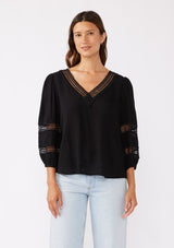 [Color: Black] A brunette model wearing a black casual top with lace trim or crochet inserts. With a v neckline, long sleeve with elastic cuff, and a flowy fit designed with crinkled rayon fabric. A summer blouse perfectly styled with denim shorts.