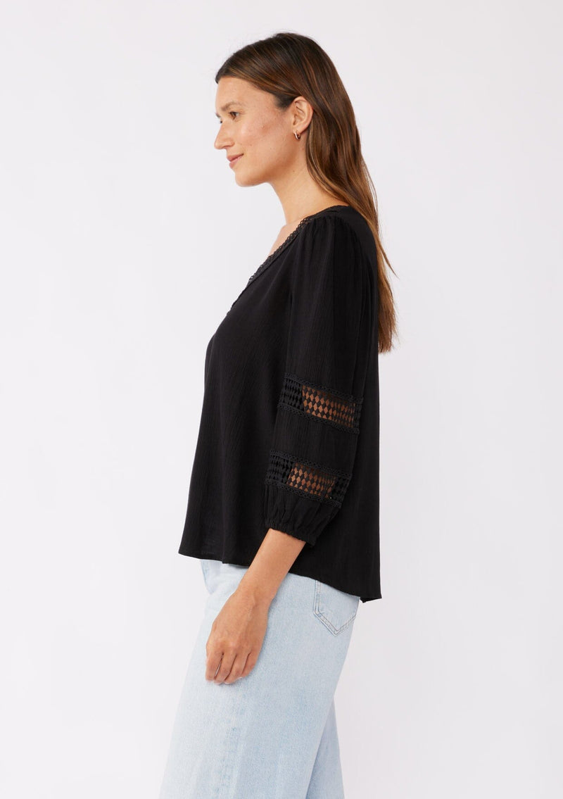 [Color: Black] A brunette model wearing a black casual top with lace trim or crochet inserts. With a v neckline, long sleeve with elastic cuff, and a flowy fit designed with crinkled rayon fabric. A summer blouse perfectly styled with denim shorts.