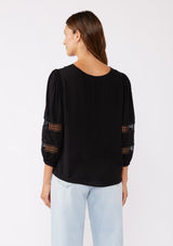 [Color: Black] A brunette model wearing a black casual top with lace trim or crochet inserts. With a v neckline, long sleeve with elastic cuff, and a flowy fit designed with crinkled rayon fabric. A summer blouse perfectly styled with denim shorts.