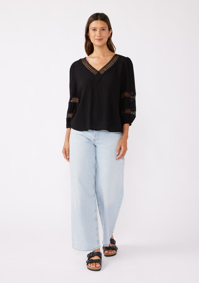 [Color: Black] A brunette model wearing a black casual top with lace trim or crochet inserts. With a v neckline, long sleeve with elastic cuff, and a flowy fit designed with crinkled rayon fabric. A summer blouse perfectly styled with denim shorts.