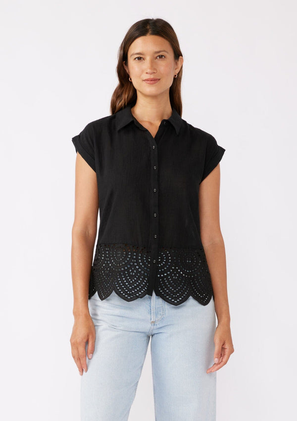 [Color: Black] A brunette model wearing a breezy cotton camp shirt with a unique embroidered eyelet design and scalloped edge. This black collared top features a functional button front, cap sleeves, and a relaxed fit. Paired with light wash denim jeans for that effortless laid back style