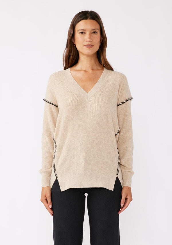 [Color: Oatmeal] A brunette model wearing an everyday sweater with a relaxed silhouette in a beige tan. With a classic v neckline, long sleeves, and an exposed seam with blanket stitch. A fall sweater styled with black bottom and casual sandals. 