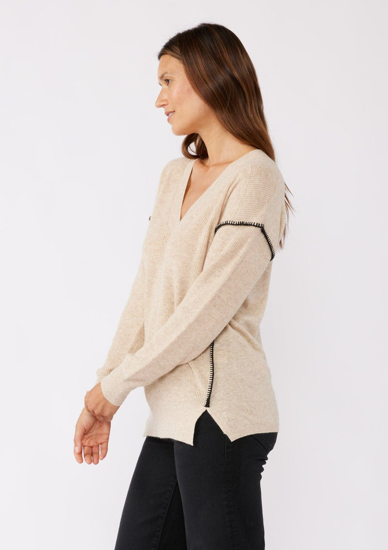 [Color: Oatmeal] A brunette model wearing an everyday sweater with a relaxed silhouette in a beige tan. With a classic v neckline, long sleeves, and an exposed seam with blanket stitch. A fall sweater styled with black bottom and casual sandals. 