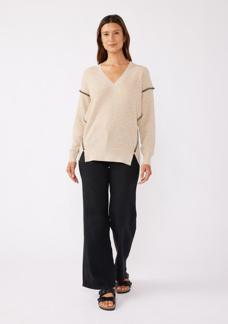 [Color: Oatmeal] A brunette model wearing an everyday sweater with a relaxed silhouette in a beige tan. With a classic v neckline, long sleeves, and an exposed seam with blanket stitch. A fall sweater styled with black bottom and casual sandals. 