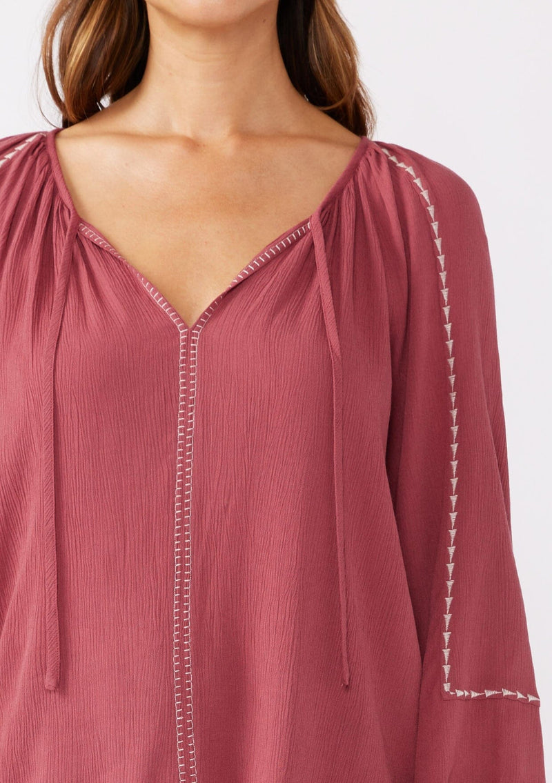 [Color: Mauve/Natural] A detailed image of a brunette model wearing an embroidered pink bohemian blouse. With a split v neckline with ties, long raglan sleeves, and an elastic cuff. A relaxed fit boho top styled with black bottoms for the ultimate fall ensemble.