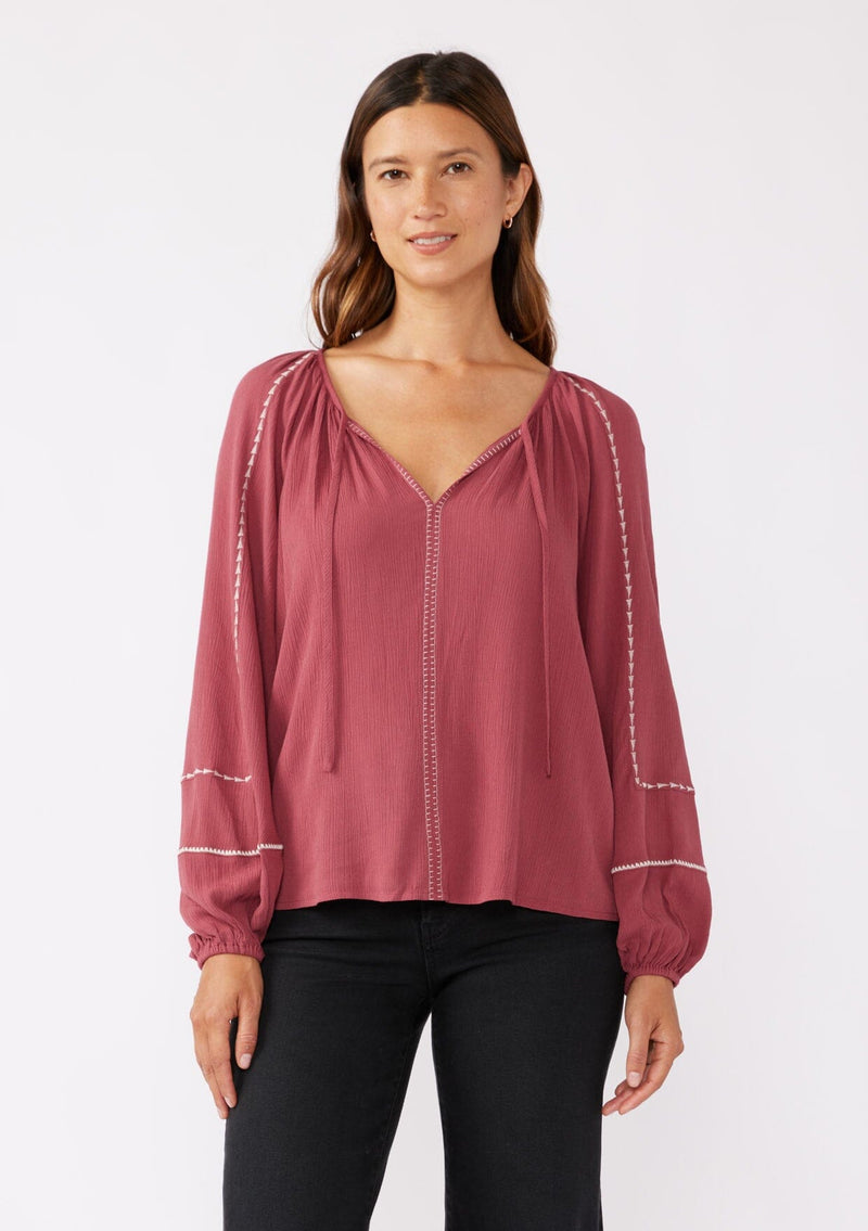 [Color: Mauve/Natural] A front image of a brunette model wearing an embroidered pink bohemian blouse. With a split v neckline with ties, long raglan sleeves, and an elastic cuff. A relaxed fit boho top styled with black bottoms for the ultimate fall ensemble.