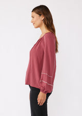 [Color: Mauve/Natural] A side image of a brunette model wearing an embroidered pink bohemian blouse. With a split v neckline with ties, long raglan sleeves, and an elastic cuff. A relaxed fit boho top styled with black bottoms for the ultimate fall ensemble.