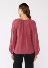 [Color: Mauve/Natural] A back image of a brunette model wearing an embroidered pink bohemian blouse. With a split v neckline with ties, long raglan sleeves, and an elastic cuff. A relaxed fit boho top styled with black bottoms for the ultimate fall ensemble.