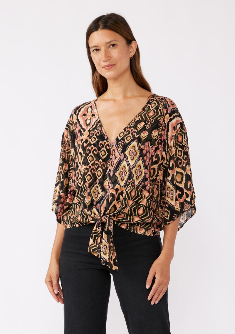 [Color: Grey/Dusty Rose] A brunette model wearing an ultra bohemian tie front top in an orange geometric print with western inspiration. Complete with a knot detail at the hem, v neckline, and flowy kimono sleeves. 