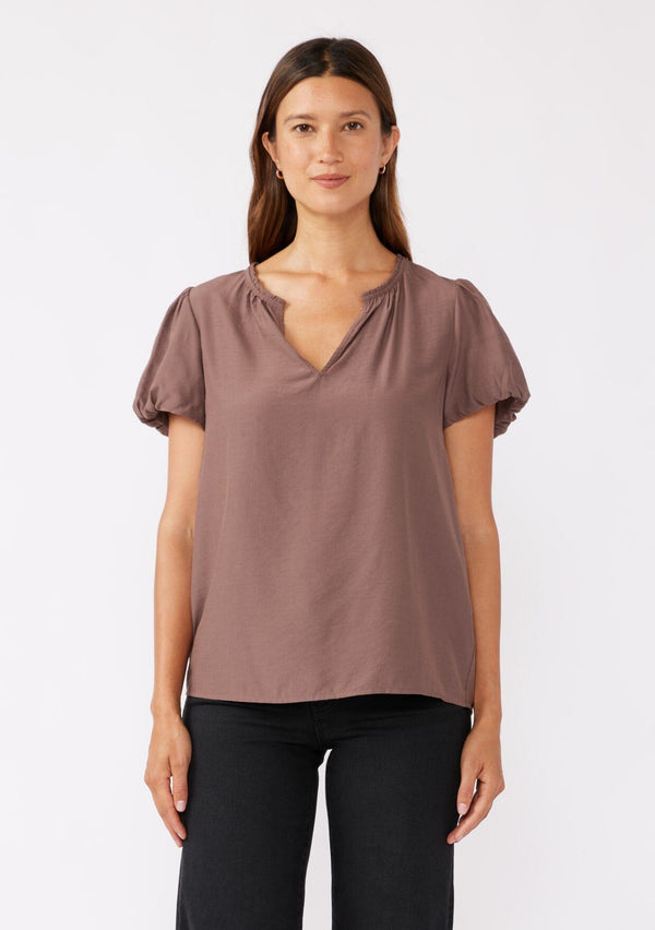 [Color: Mocha] A brunette model wearing a sophisticated classic brown top with black denim jeans. Featuring bubble puff sleeves, ruffled neckline, and a tie neck detail. A relaxed fit top perfect for the office or as a formal attire.