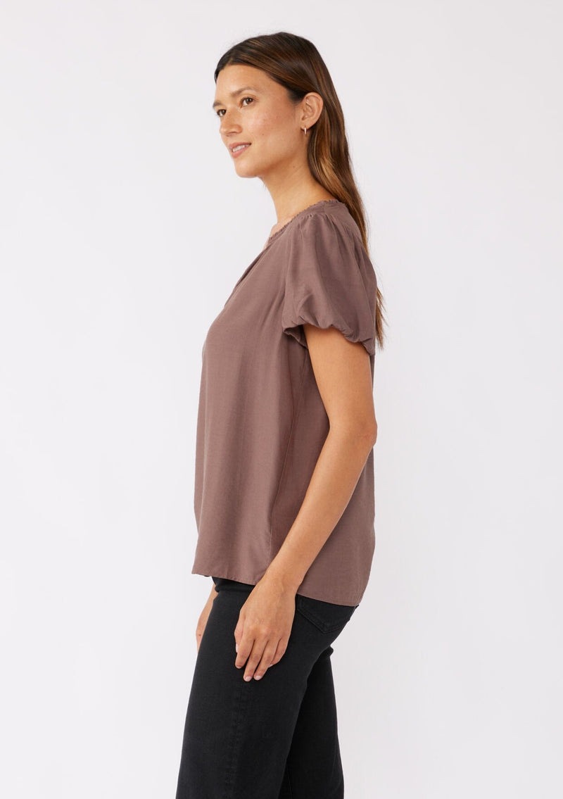 [Color: Mocha] A brunette model wearing a sophisticated classic brown top with black denim jeans. Featuring bubble puff sleeves, ruffled neckline, and a tie neck detail. A relaxed fit top perfect for the office or as a formal attire.