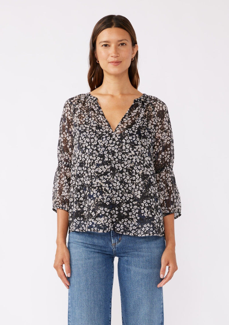 [Color: Black/Grey] A brunette woman wearing an ultra flowy chiffon blouse with metallic clip dot details. A black floral blouse with a split v neckline, long sleeves, and elastic cuffs. Paired with denim jeans for a relaxed fall look, perfect for dinner and classy evening parties.  