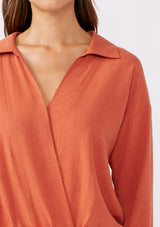 [Color: Spice] A brunette model wearing an orange blouse in a soft linen blend. With a collared neckline, flattering v neckline, front hook and eye closure, and long sleeves with button cuffs. Completed with a comfortable half elastic waist at the back for a comfortable fit. Styled with a wide leg denim for casual laid back look.