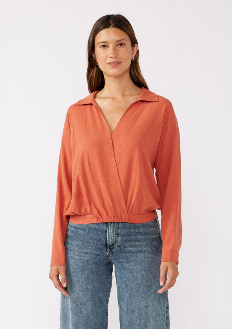 [Color: Spice] A brunette model wearing an orange blouse in a soft linen blend. With a collared neckline, flattering v neckline, front hook and eye closure, and long sleeves with button cuffs. Completed with a comfortable half elastic waist at the back for a comfortable fit. Styled with a wide leg denim for casual laid back look.