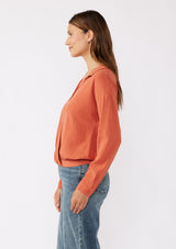 [Color: Spice] A brunette model wearing an orange blouse in a soft linen blend. With a collared neckline, flattering v neckline, front hook and eye closure, and long sleeves with button cuffs. Completed with a comfortable half elastic waist at the back for a comfortable fit. Styled with a wide leg denim for casual laid back look.