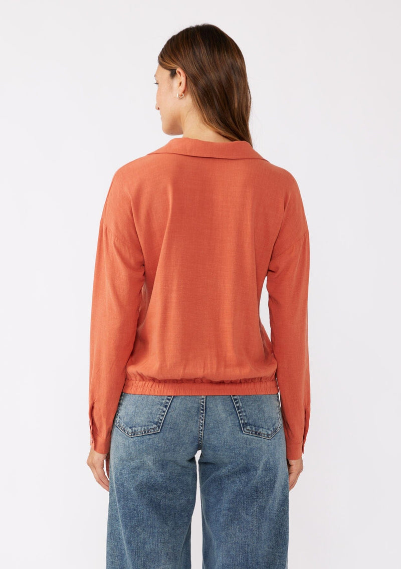 [Color: Spice] A brunette model wearing an orange blouse in a soft linen blend. With a collared neckline, flattering v neckline, front hook and eye closure, and long sleeves with button cuffs. Completed with a comfortable half elastic waist at the back for a comfortable fit. Styled with a wide leg denim for casual laid back look.