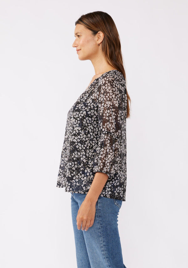 [Color: Black/Grey] A brunette woman wearing an ultra flowy chiffon blouse with metallic clip dot details. A black floral blouse with a split v neckline, long sleeves, and elastic cuffs. Paired with denim jeans for a relaxed fall look, perfect for dinner and classy evening parties.