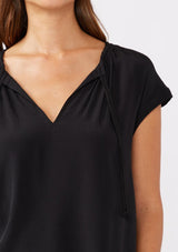 [Color: Black] A brunette model wearing a relaxed fit black top. With self neck ties, ruched split v neckline, and short dolman sleeves. Styled with a cute denim bottoms for a laid back look.