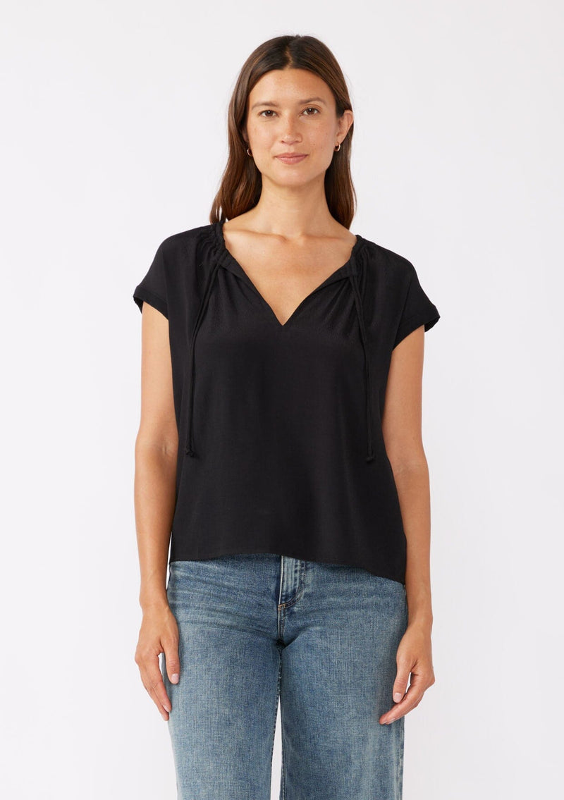 [Color: Black] A brunette model wearing a relaxed fit  black top. With self neck ties, ruched split v neckline, and short dolman sleeves. Styled with a cute denim bottoms for a laid back look. 