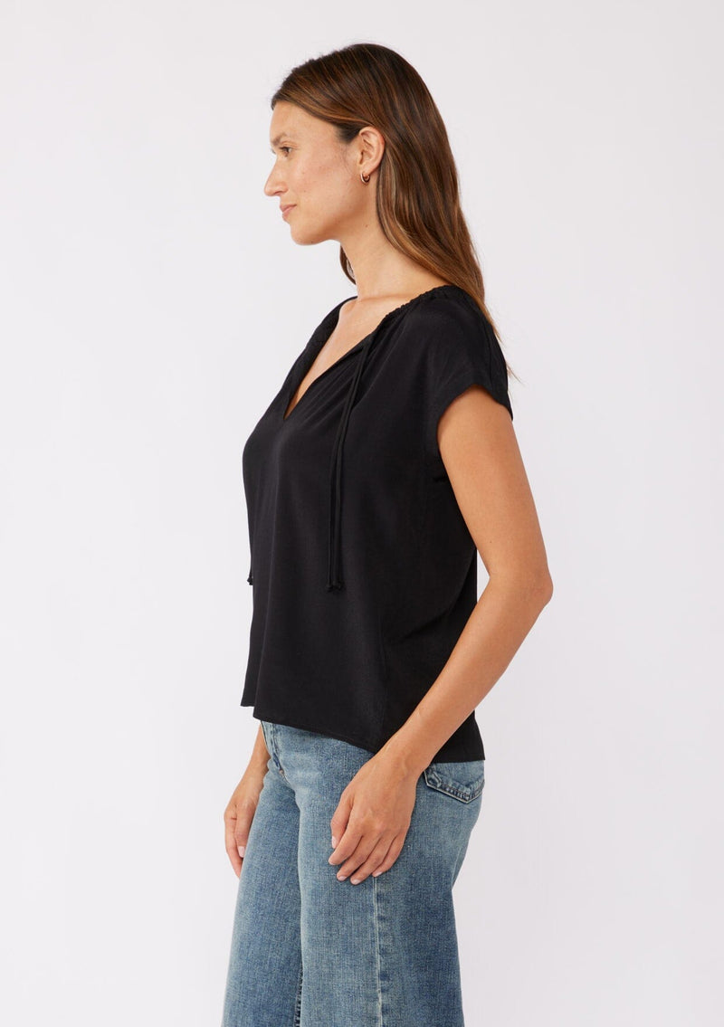 [Color: Black] A brunette model wearing a relaxed fit black top. With self neck ties, ruched split v neckline, and short dolman sleeves. Styled with a cute denim bottoms for a laid back look.