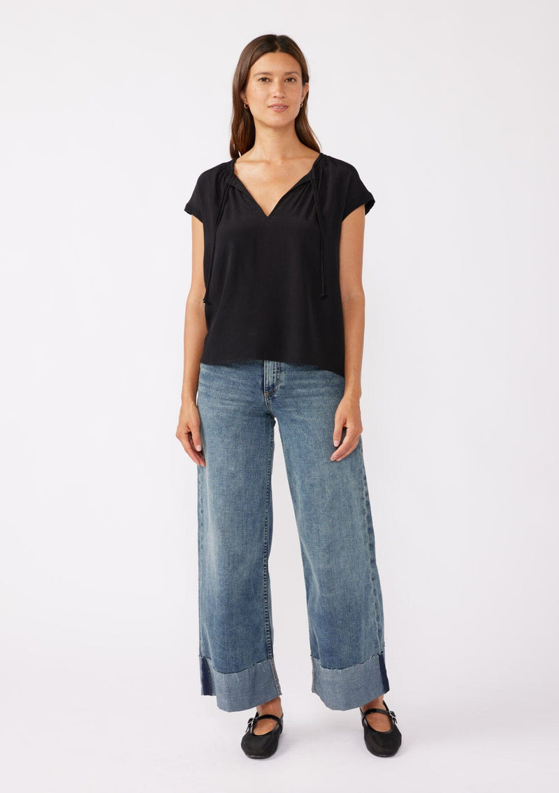 [Color: Black] A brunette model wearing a relaxed fit black top. With self neck ties, ruched split v neckline, and short dolman sleeves. Styled with a cute denim bottoms for a laid back look.