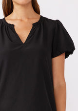 [Color: Black] A brunette model wearing a sophisticated classic black top with denim jeans. Featuring bubble puff sleeves, ruffled neckline, and a tie neck detail. A relaxed fit top perfect for the office or as a formal attire.