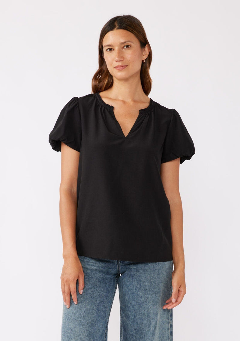 [Color: Black] A brunette model wearing a sophisticated classic black top with denim jeans. Featuring bubble puff sleeves, ruffled neckline, and a tie neck detail. A relaxed fit top perfect for the office or as a formal attire.