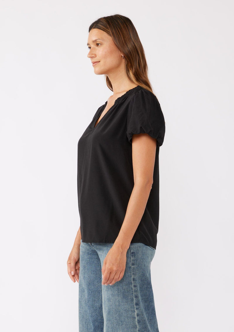 [Color: Black] A brunette model wearing a sophisticated classic black top with denim jeans. Featuring bubble puff sleeves, ruffled neckline, and a tie neck detail. A relaxed fit top perfect for the office or as a formal attire.