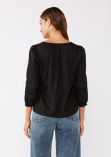 [Color: Black] A brunette model wearing a bohemian black blouse with embroidered lace details. Crafted from a rayon and linen blend, this top feature long sleeves, a lace up front, and elastic cuffs. Perfect addition to any fall bohemian wardrobe.  