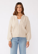 [Color: Natural] A brunette model wearing an off white, ivory cozy knit sweater with a zipper front. Featuring long sleeves and classic ribbed trims. A versatile sweater perfect for layering and styling any fall ensemble. 