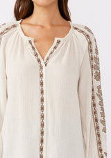 [Color: Natural/Mocha] A brunette model wearing an ultra bohemian blouse in off white with brown embroidery. Featuring a split v neckline, long sleeves, and a relaxed fit.