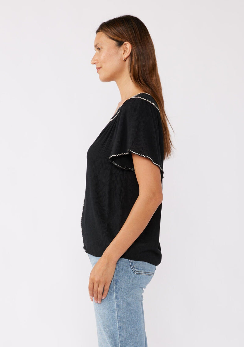 [Color: Black/Natural] A brunette model wearing a classic bohemian top in black. With an embroidered blanket stitch trim, short flutter sleeves, and a split v neckline. Perfect for any fall wardrobe; styled with denim for a cool and casual look.  