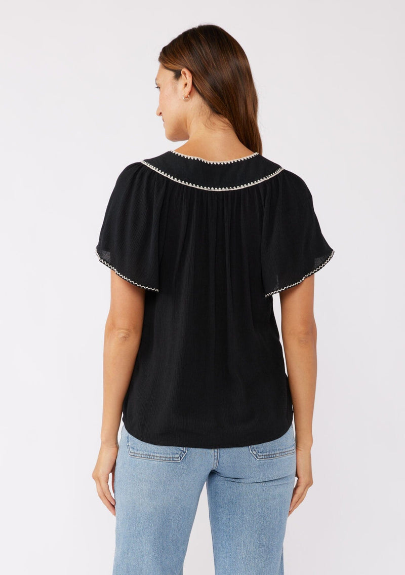 [Color: Black/Natural] A brunette model wearing a classic bohemian top in black. With an embroidered blanket stitch trim, short flutter sleeves, and a split v neckline. Perfect for any fall wardrobe; styled with denim for a cool and casual look.  