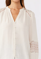 [Color: Natural/Taupe] A brunette model wearing an off white bohemian blouse with embroidered details. Featuring a split v neckline, self covered button front, and long sleeves with smocked cuffs.