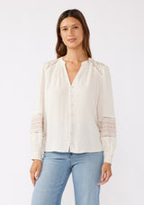 [Color: Natural/Taupe] A brunette model wearing an off white bohemian blouse with embroidered details. Featuring a split v neckline, self covered button front, and long sleeves with smocked cuffs. Paired with light wash denim for the perfect fall look. 