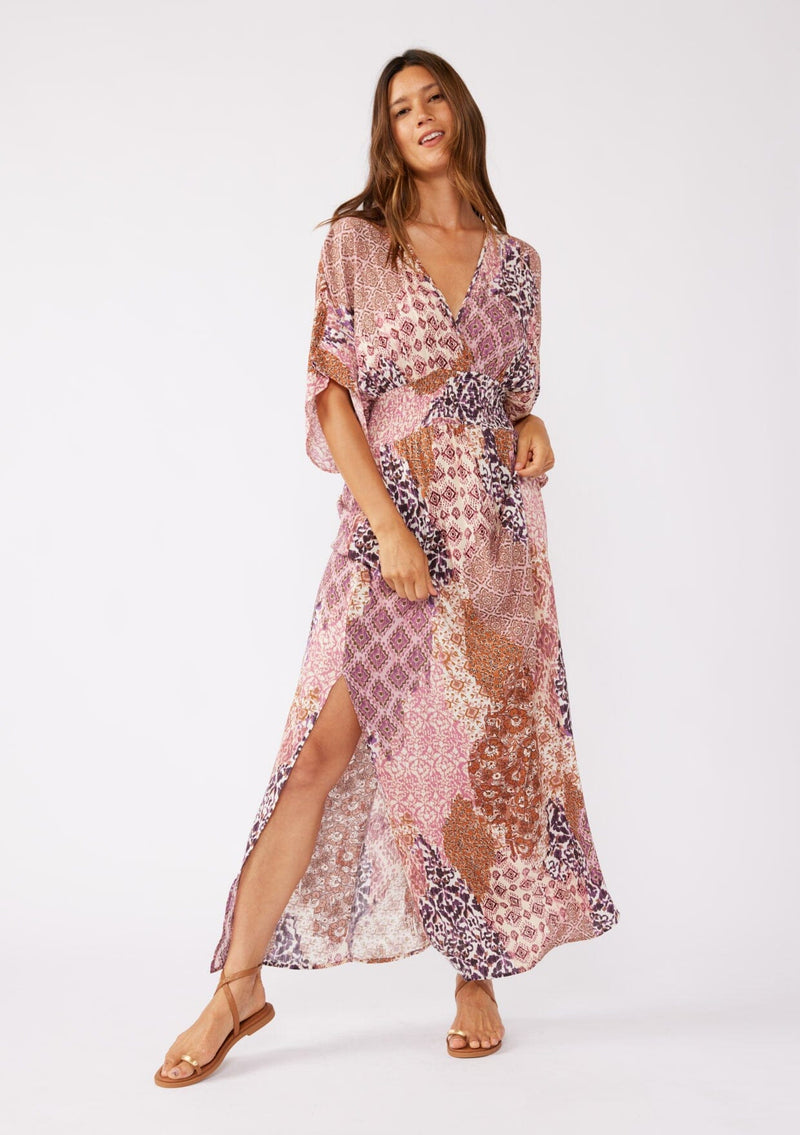 [Color: Natural/Clay] A blonde model wearing an ultra bohemian maxi dress in a pink patchwork print. Featuring flowy kimono sleeves, v neckline, smocked waist, a sexy open back with tie detail, and side slits. Perfect as a beach cover up or casual outings. 