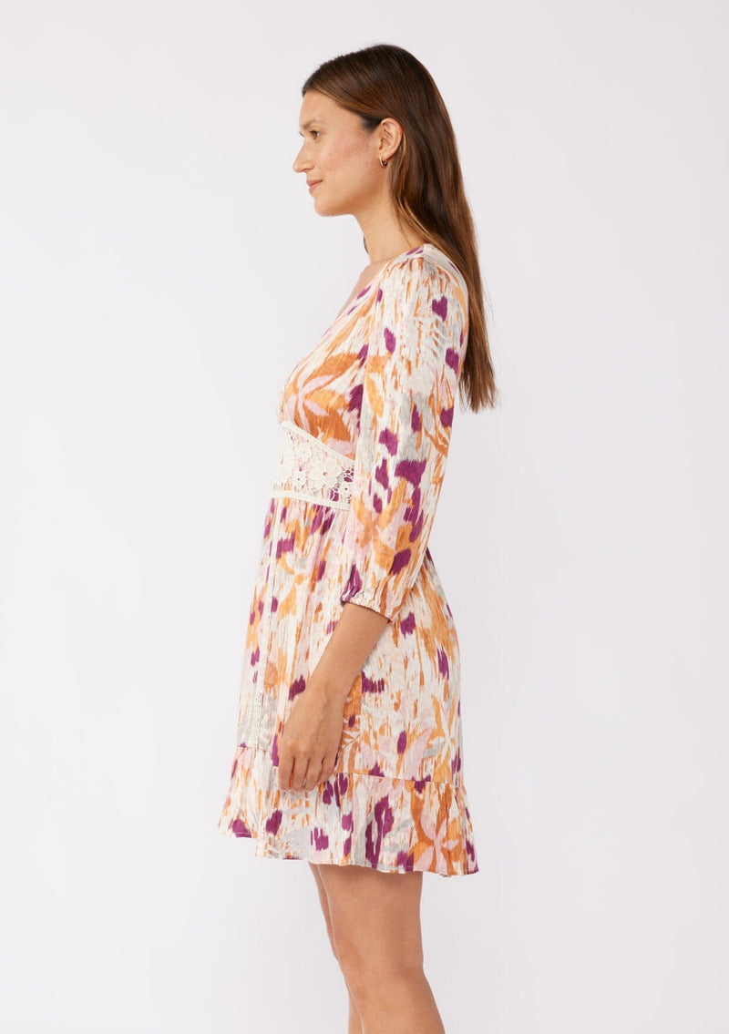 [Color: Natural/Plum] A brunette model wearing a bohemian mini dress in an orange and purple abstract ikat print. Featuring ¾ length sleeves, a v-neckline, ruffle trimmed skirt, and floral lace details. A relaxed fit style designed for the Fall season. 