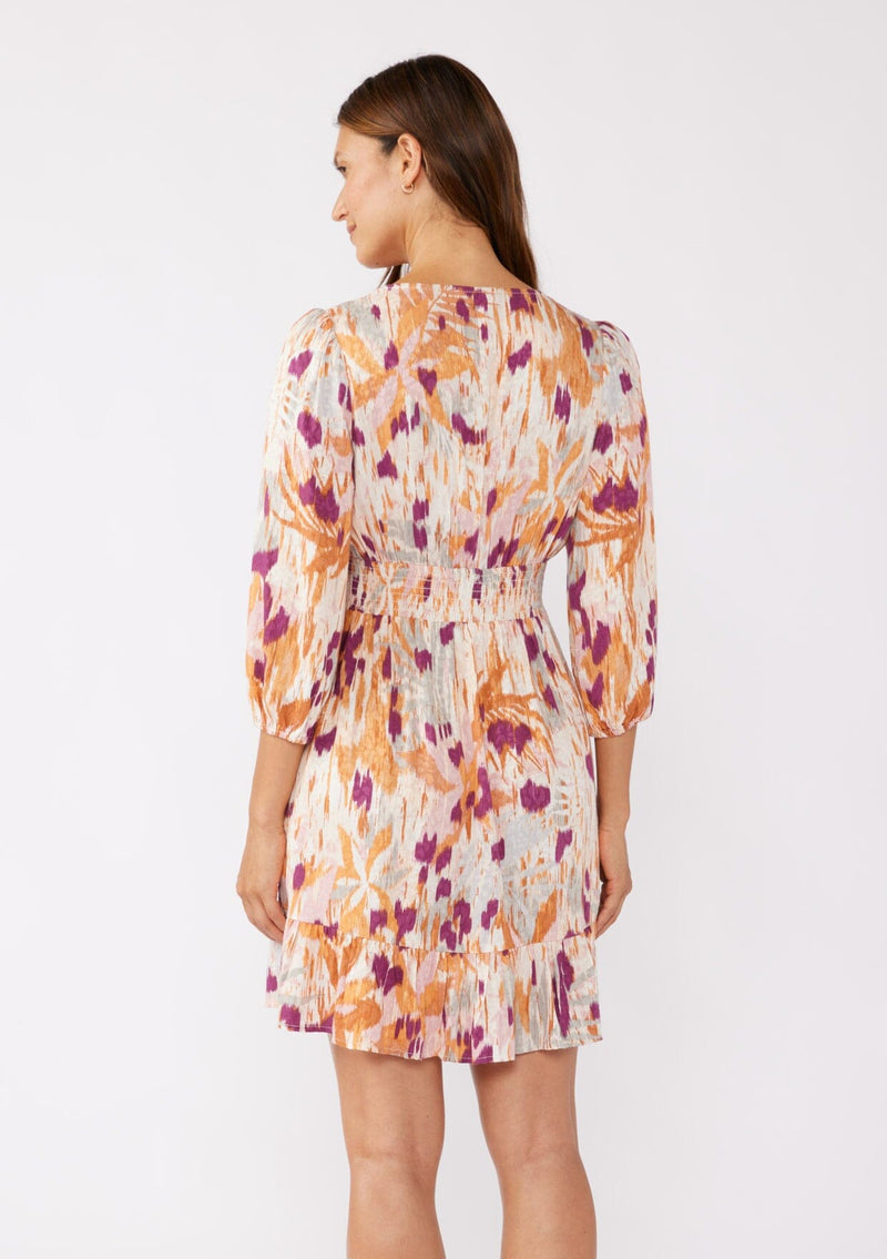 [Color: Natural/Plum] A back image of a brunette model wearing a bohemian mini dress in an orange and purple abstract ikat print. Featuring ¾ length sleeves, a v-neckline, ruffle trimmed skirt, and floral lace details. A relaxed fit style designed for the Fall season. 