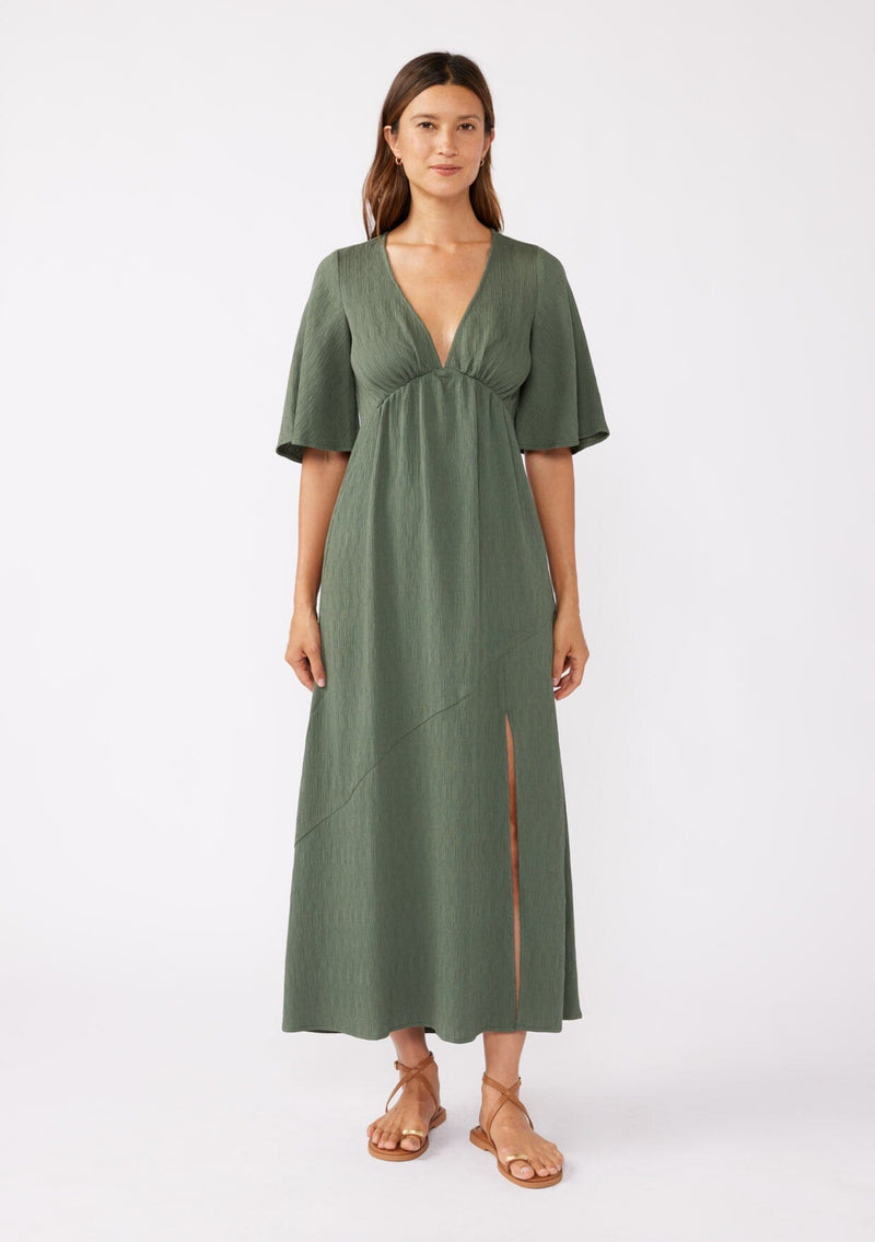 [Color: Olive] A brunette model wearing a textured maxi dress in a green autumn hue. With a deep v neckline, empire waistline, flowy half sleeves, and a side slit. Paired with casual sandals for any daytime outing. 