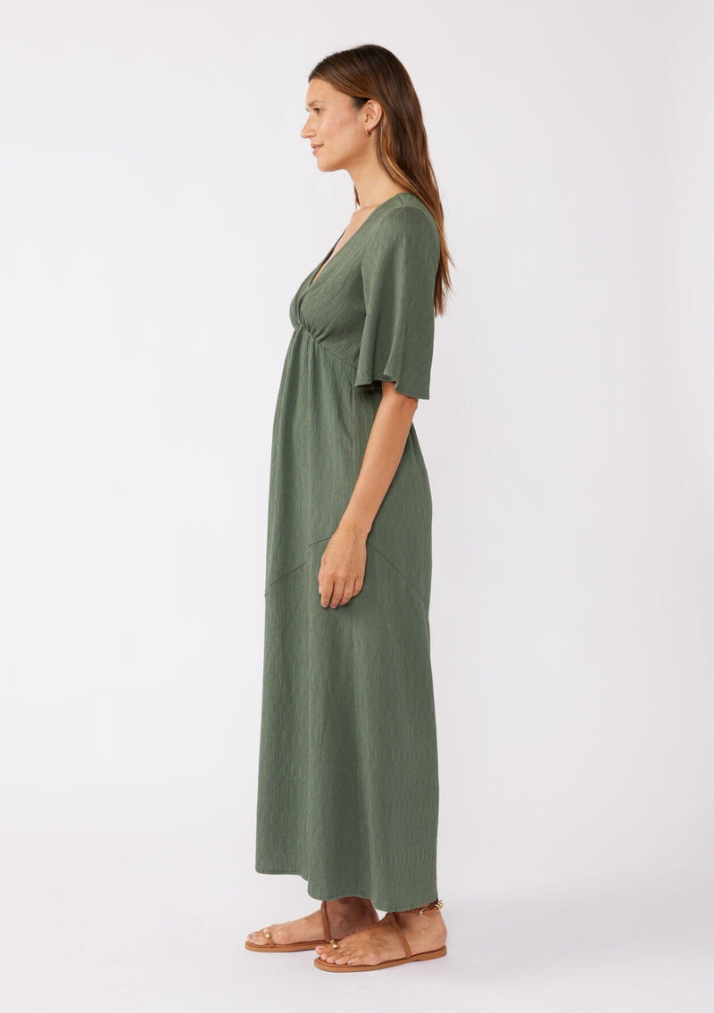 [Color: Olive] A brunette model wearing a textured maxi dress in a green autumn hue. With a deep v neckline, empire waistline, flowy half sleeves, and a side slit. Paired with casual sandals for any daytime outing.