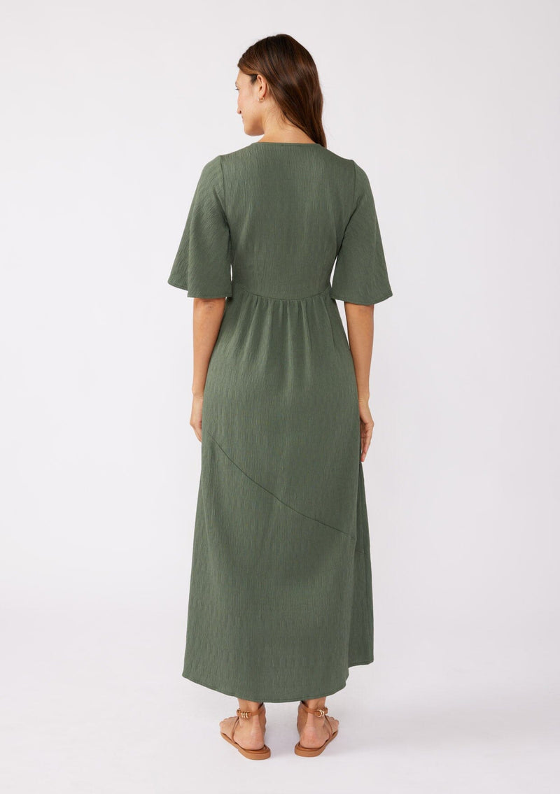 [Color: Olive] A brunette model wearing a textured maxi dress in a green autumn hue. With a deep v neckline, empire waistline, flowy half sleeves, and a side slit. Paired with casual sandals for any daytime outing.