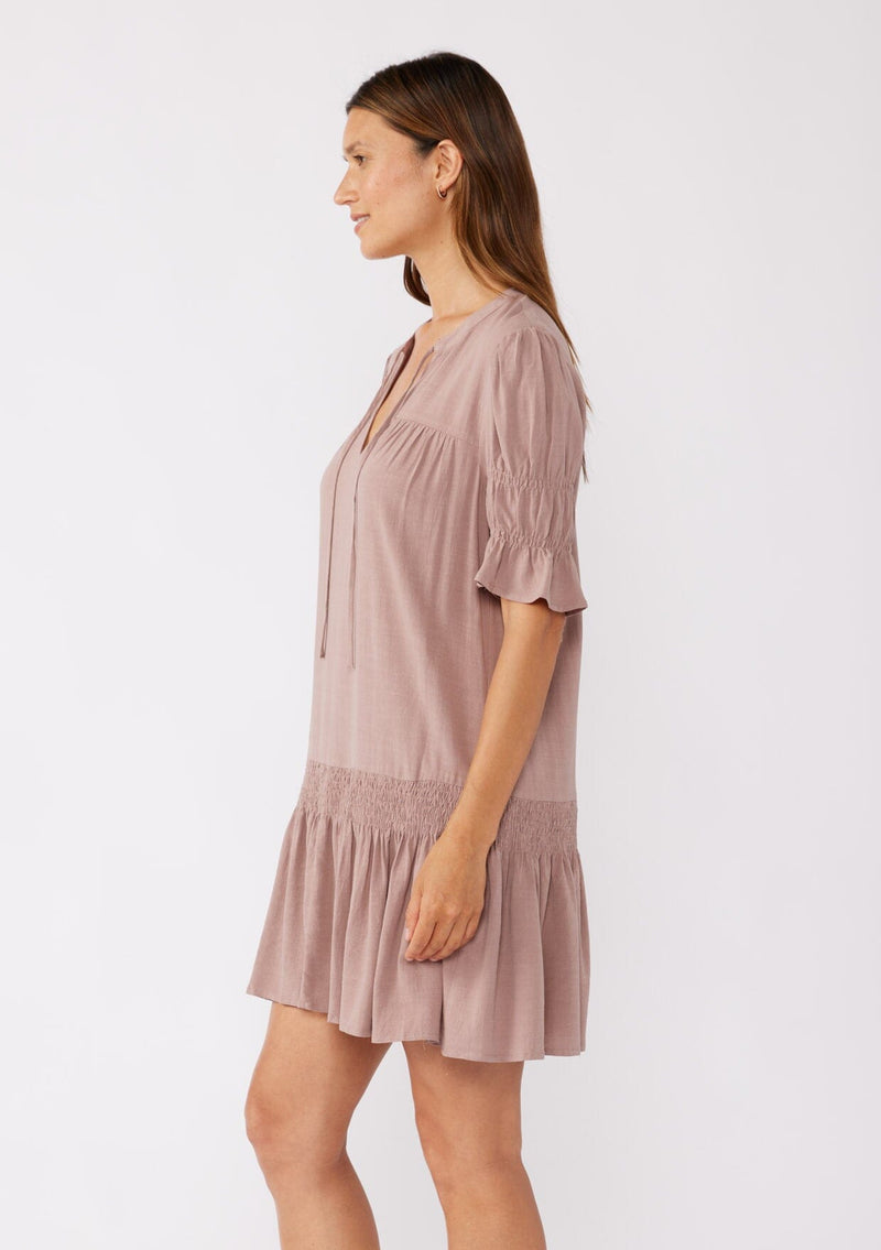 [Color: Light Mocha] A side facing image of a brunette model wearing a pink, ultra-flowy bohemian mini dress with a drop waist. Featuring short puff sleeves, a split v-neckline with tie detail, and a smocked tiered skirt. A casual fall mini dress in a baby doll silhouette crafted in rayon and linen blend.  