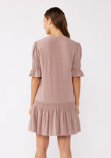 [Color: Light Mocha] A back facing image of a brunette model wearing a pink, ultra-flowy bohemian mini dress with a drop waist. Featuring short puff sleeves, a split v-neckline with tie detail, and a smocked tiered skirt. A casual fall mini dress in a baby doll silhouette crafted in rayon and linen blend.  