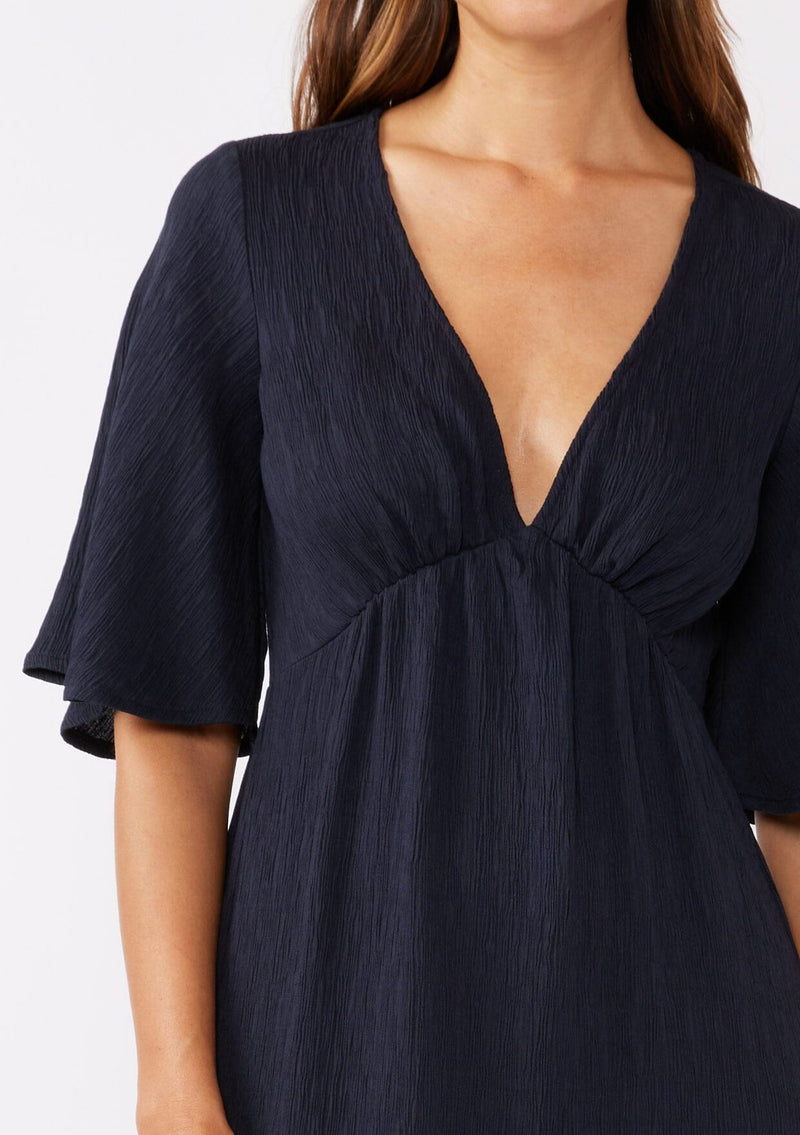 [Color: Navy] A brunette model wearing a textured maxi dress in a deep navy blue. With a deep v neckline, empire waistline, flowy half sleeves, and a side slit. Paired with casual sandals for any daytime outing.