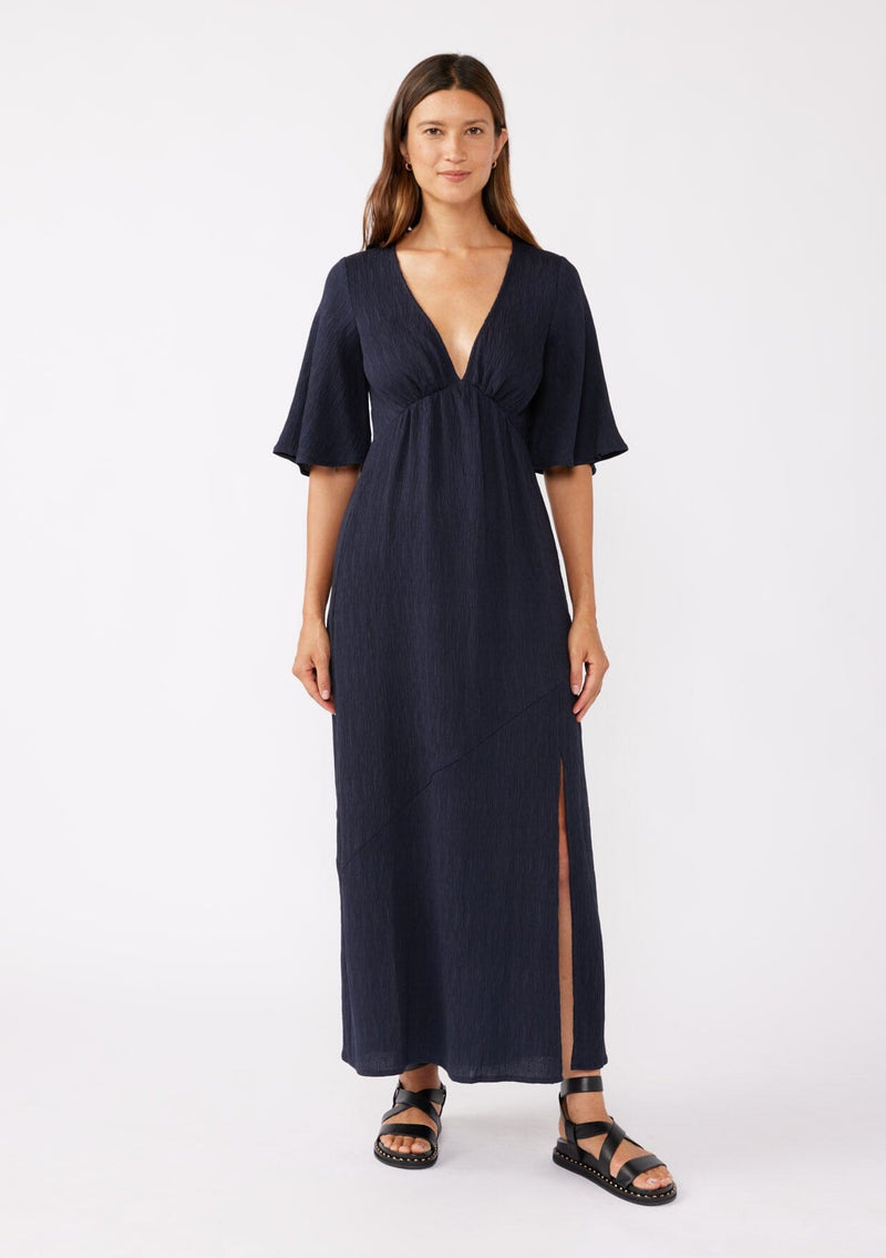 [Color: Navy] A brunette model wearing a textured maxi dress in a deep navy blue. With a deep v neckline, empire waistline, flowy half sleeves, and a side slit. Paired with casual sandals for any daytime outing.