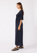 [Color: Navy] A brunette model wearing a textured maxi dress in a deep navy blue. With a deep v neckline, empire waistline, flowy half sleeves, and a side slit. Paired with casual sandals for any daytime outing.